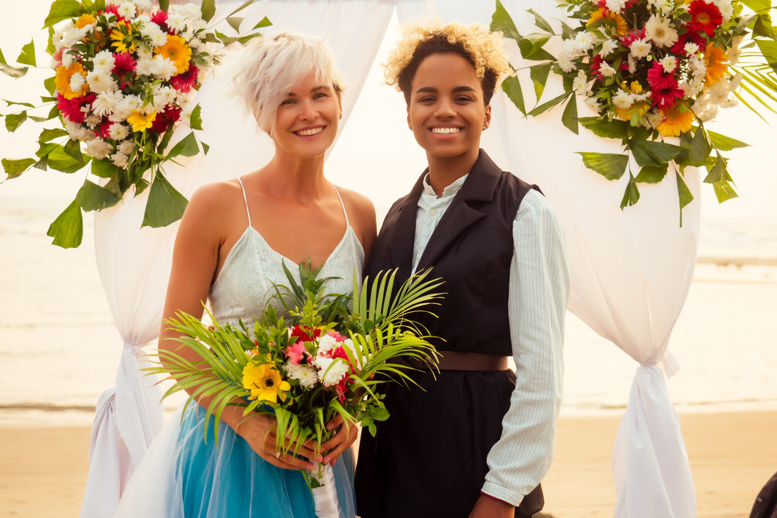 afro american female in the role of the groom and short blonde haired bride in blue dress in ceremony under wedding arch in tropical beach